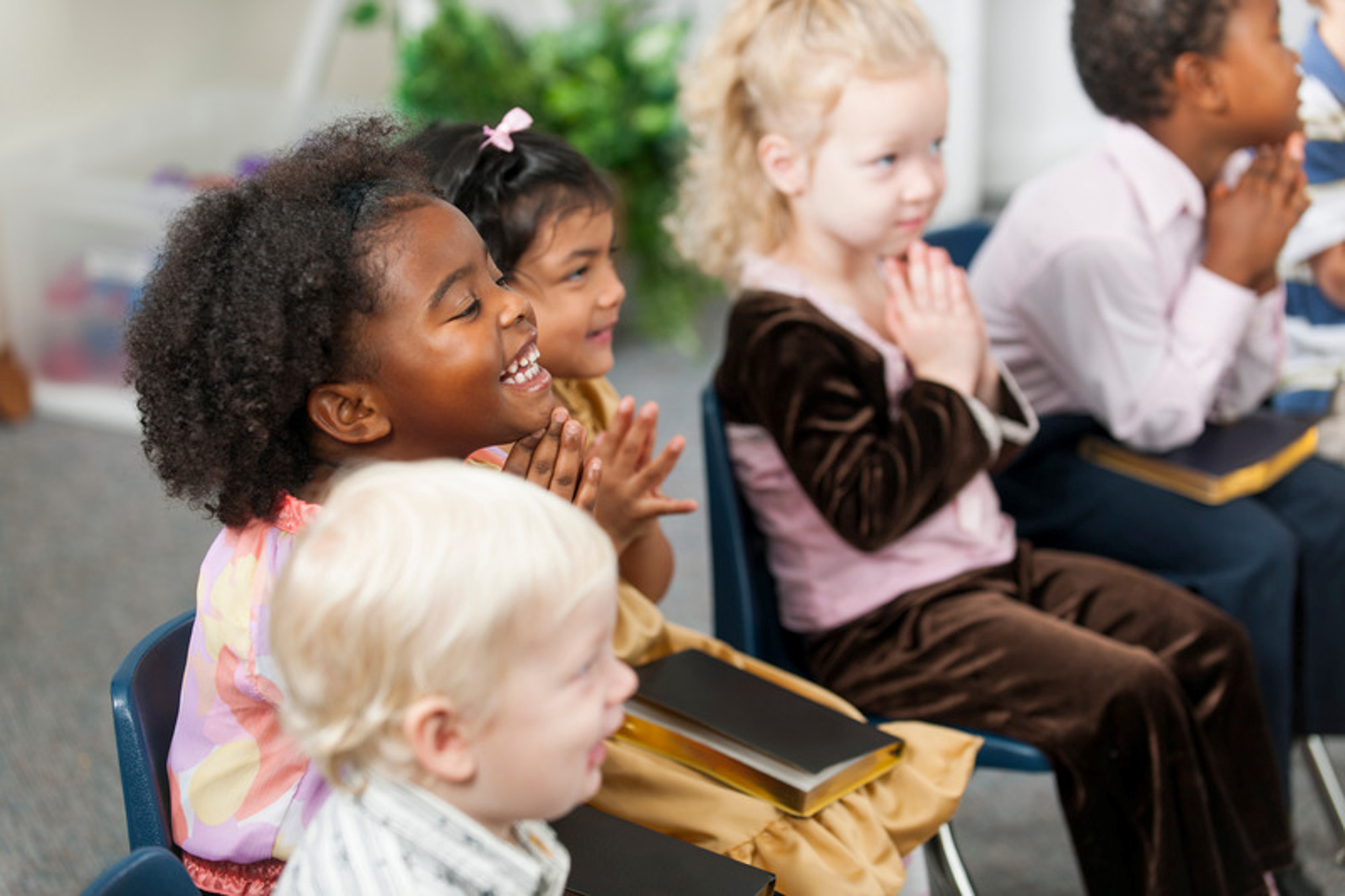 A group of young children sitting in chairs, smiling and clapping their hands, holding books in their laps in a classroom setting.
